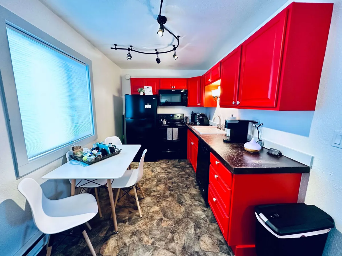 This image centers on the kitchen area, featuring vibrant red cabinets, sleek black appliances, a white dining table, and a window with blinds that brings in a cheerful touch of natural light. The scene suggests an inviting atmosphere for both cooking and dining, with visible space for meal prep and informal gatherings.