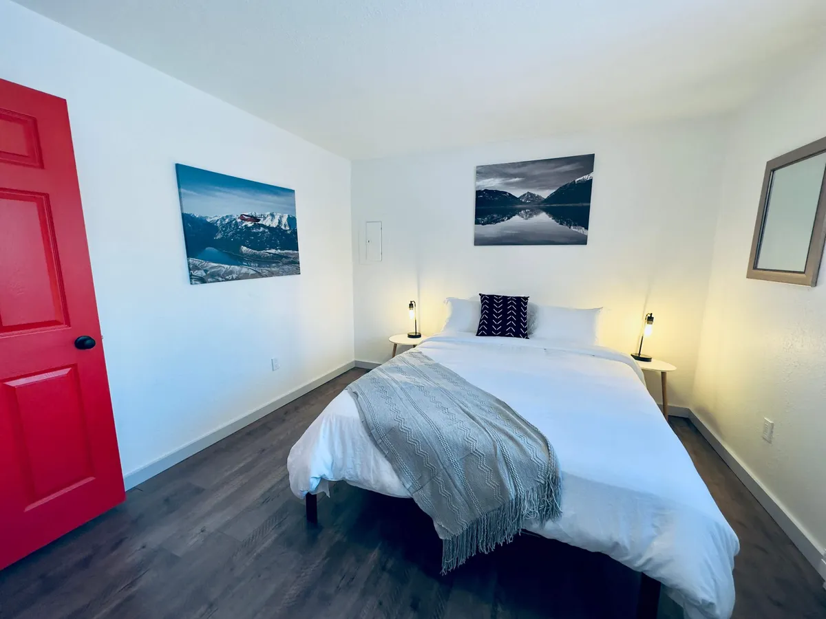 A cozy, serene corner featuring a bed with crisp white linens, a red door, and nature-inspired wall art. The arrangement creates a peaceful environment that could easily double as a quiet, private workspace for creative or focused tasks.