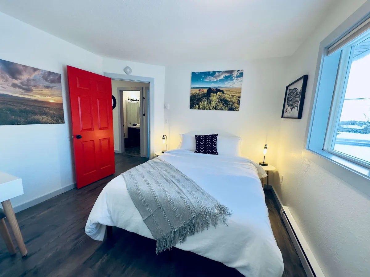 A cozy bedroom is shown, featuring a red door, a comfortable bed with decorative pillows, and wall art that adds a personal touch. Natural light streams in, creating a serene and restful environment, perfect for unwinding after outdoor activities.