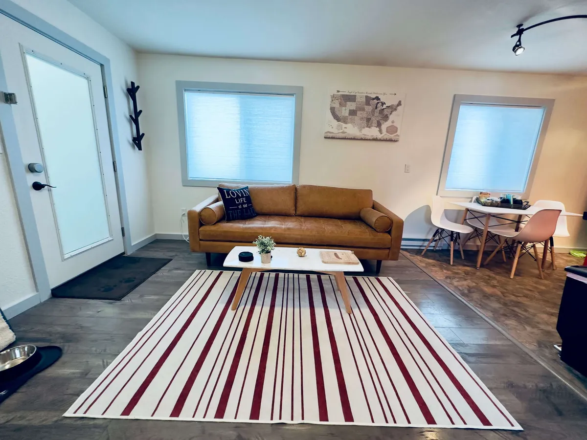 Featuring a cozy brown sofa, a striped rug, and a dining table in the background, this image offers another perspective on the dining nook area. The warm tones and soft textures create a welcoming environment ideal for socializing, relaxing, and enjoying meals together.