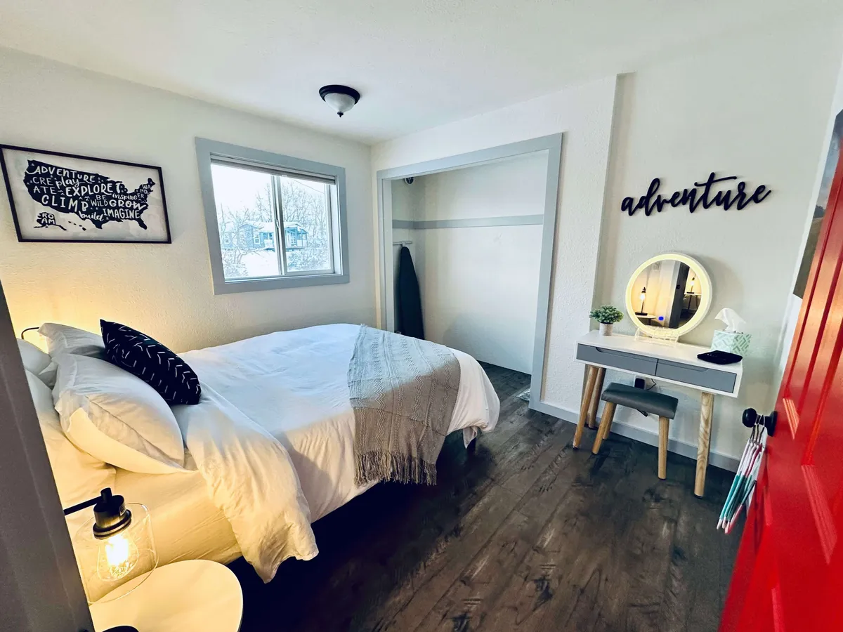 This image showcases a bedroom with crisp white linens on the bed, adventure-themed wall decor, a round mirror on the vanity, and a bright red door. The room is designed to feel inspiring and cozy, with both functional and decorative touches that invite relaxation.