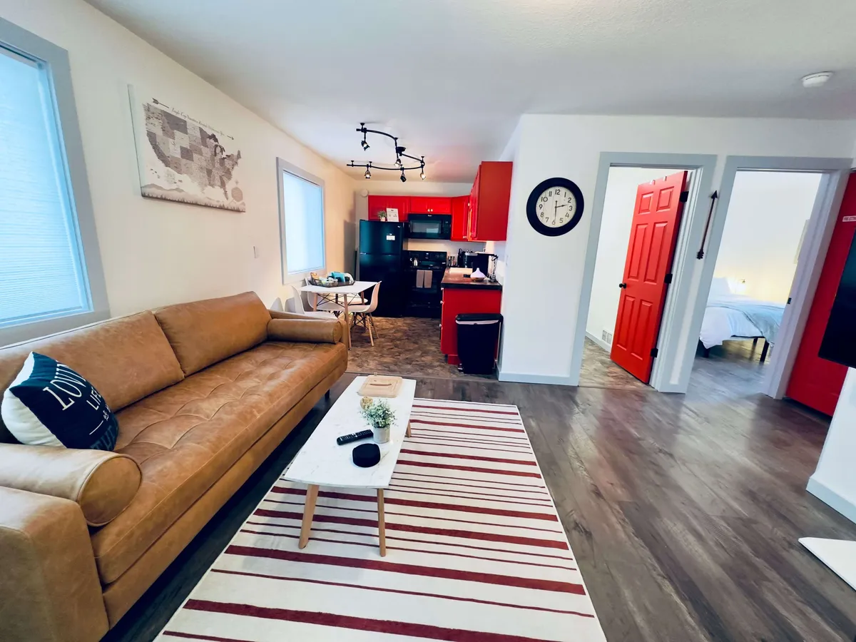 This interior shot features a sleek leather sofa, a striped area rug, red kitchen cabinets, and a dining table with chairs. The arrangement creates a seamless flow between lounge and dining areas, offering ample space to relax or connect with family and friends. Light streams in, contributing to the warm and inviting mood.