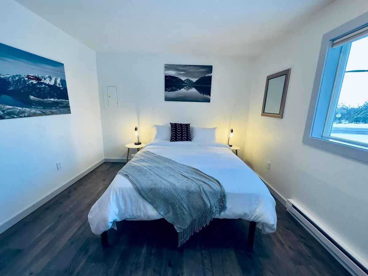 This image presents a tranquil bedroom with a bed dressed in white linens, mountain landscape artwork above the headboard, and side tables with soft-lit lamps. The setting exudes a peaceful, cozy ambiance ideal for restful sleep.