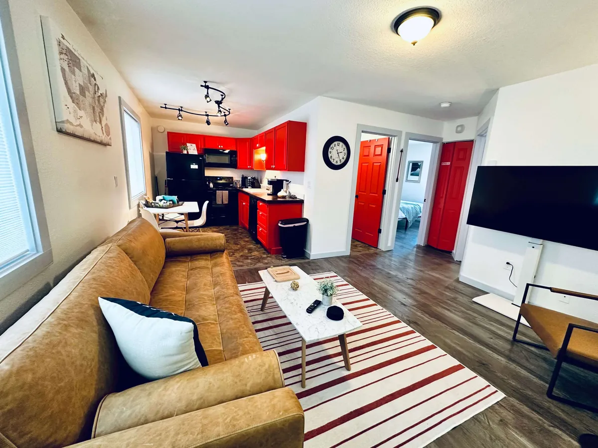 The hero image showcases a warm, contemporary living space featuring a plush brown leather sofa, vibrant red kitchen cabinets, a stylish striped area rug, and a TV on a stand—creating an inviting atmosphere for relaxation, cooking, and entertainment.