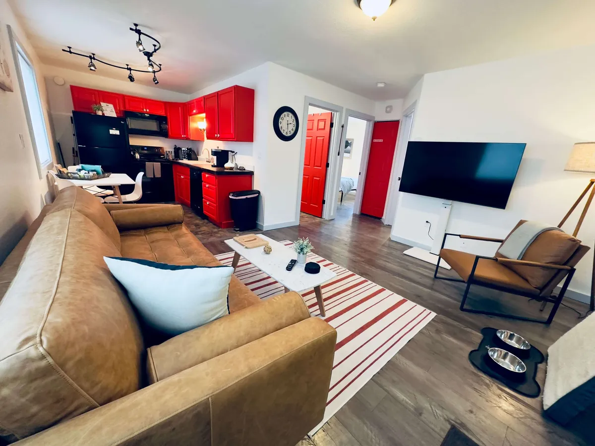 This image showcases a spacious and lively living area with a plush sofa, a large TV, red kitchen cabinets, a dining table, and a decorative rug. Natural light filters through the windows, brightening the space and inviting relaxation. The arrangement encourages both restful downtime and social gatherings, making it the central hub of the home.